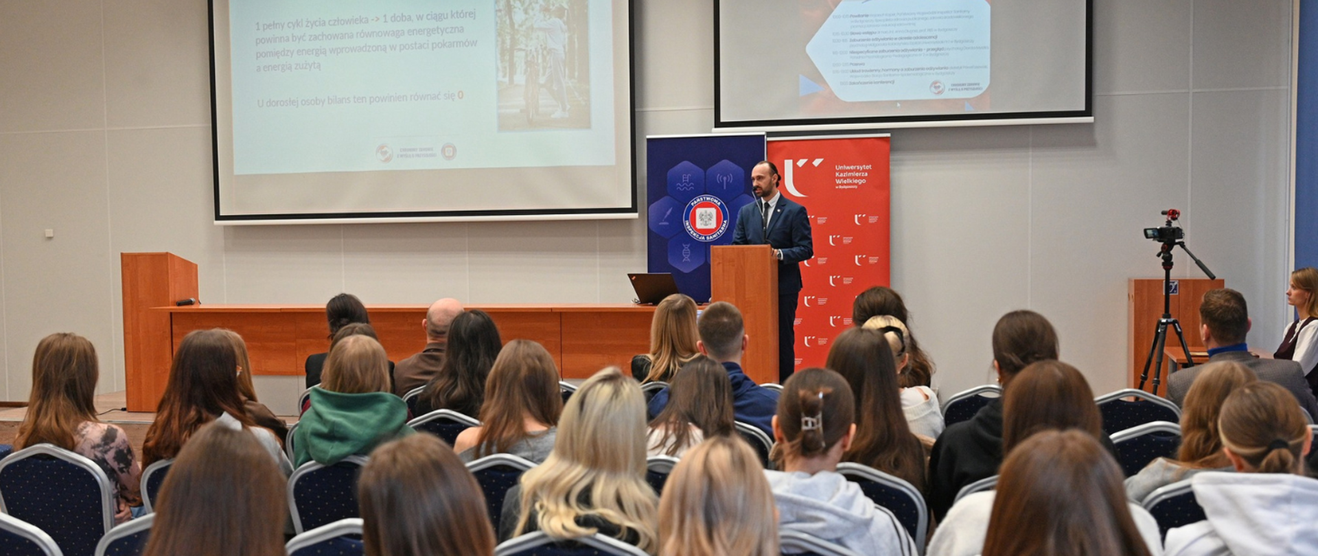 Konferencja o zaburzeniach odżywiania w Bibliotece Uniwersytetu Kazimierza Wielkiego w Bydgoszczy – na scenie stoi prelegent, a na widowni siedzą studenci oraz pracownicy Państwowej Inspekcji Sanitarnej”.