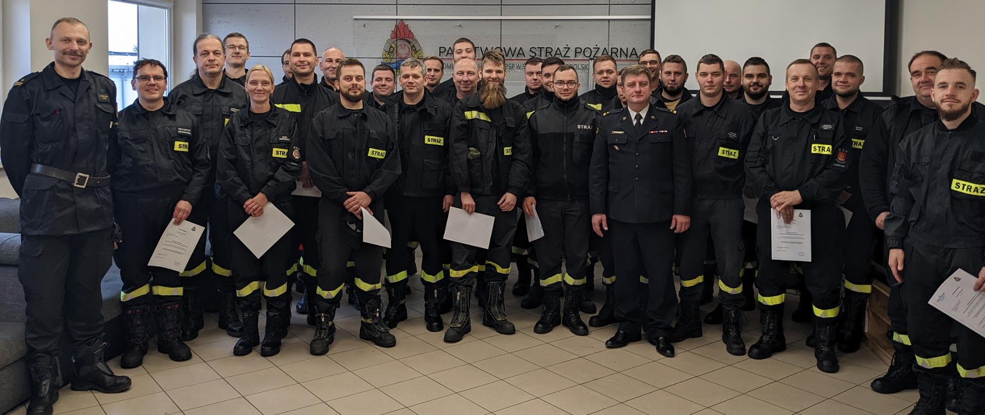 Pamiątkowa fotografia. Absolwenci szkolenia kierujących działaniem ratowniczym dla OSP stoją w rzędach trzymając zaświadczenia poświadczających ukończenie kursu.