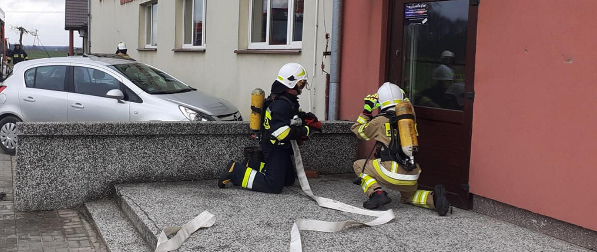 Strażacy czekają przed wejściem do budynku z linią gaśniczą.