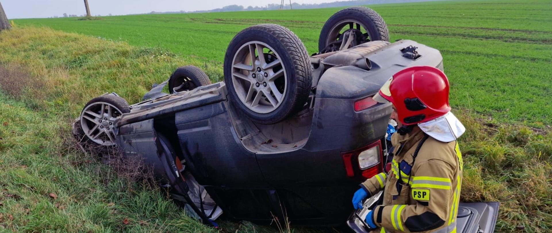 Widać uszkodzony samochód po dachowaniu od tylu, drzwi są otwarte, pojazd w rowie, po prawej jest strażak