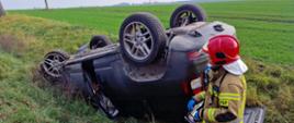 Widać uszkodzony samochód po dachowaniu od tylu, drzwi są otwarte, pojazd w rowie, po prawej jest strażak