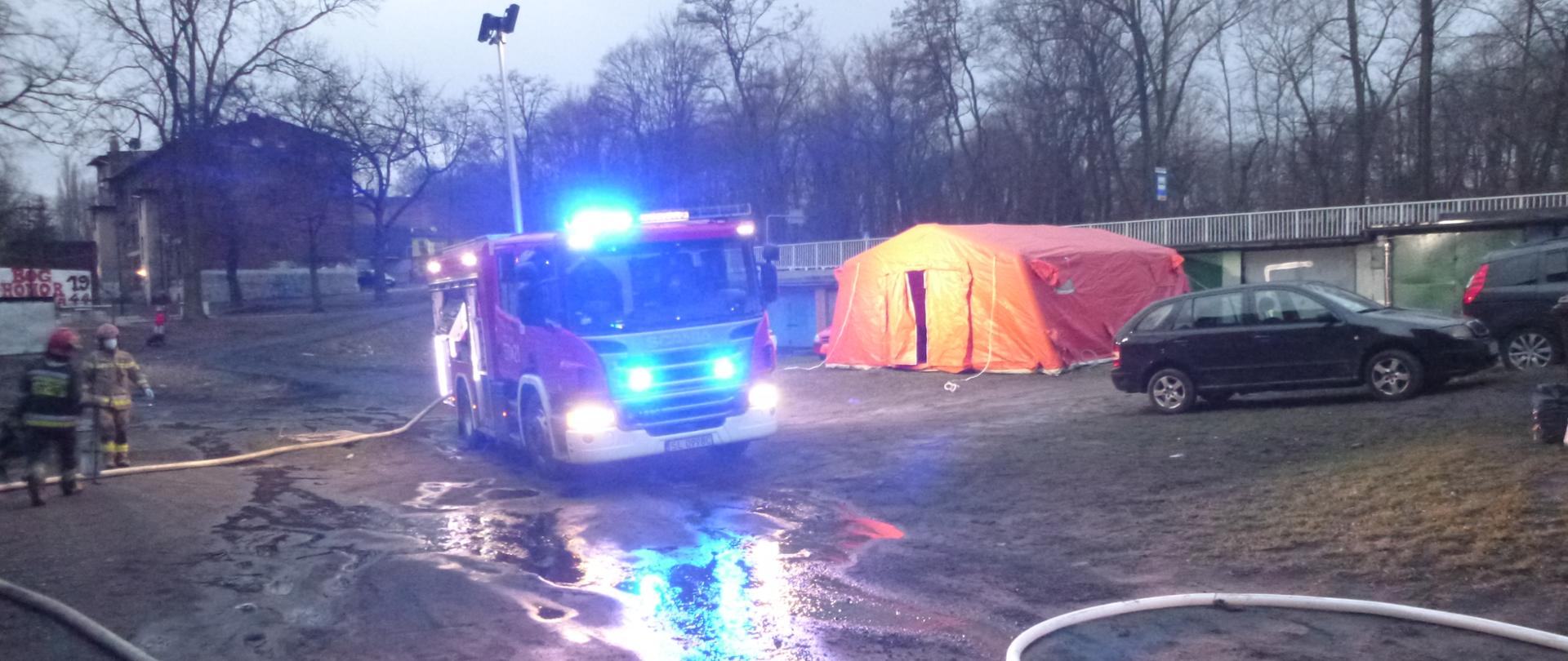 Na zdjęciu znajduje się samochód ratowniczo gaśniczy z załączonymi sygnałami świetlnymi oraz rozłożonym masztem oświetleniowym, który oświetla teren obok rozłożonego namiotu pneumatycznego koloru pomarańczowego, po lewej stronie dwóch strażaków. 