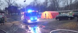 Na zdjęciu znajduje się samochód ratowniczo gaśniczy z załączonymi sygnałami świetlnymi oraz rozłożonym masztem oświetleniowym, który oświetla teren obok rozłożonego namiotu pneumatycznego koloru pomarańczowego, po lewej stronie dwóch strażaków. 