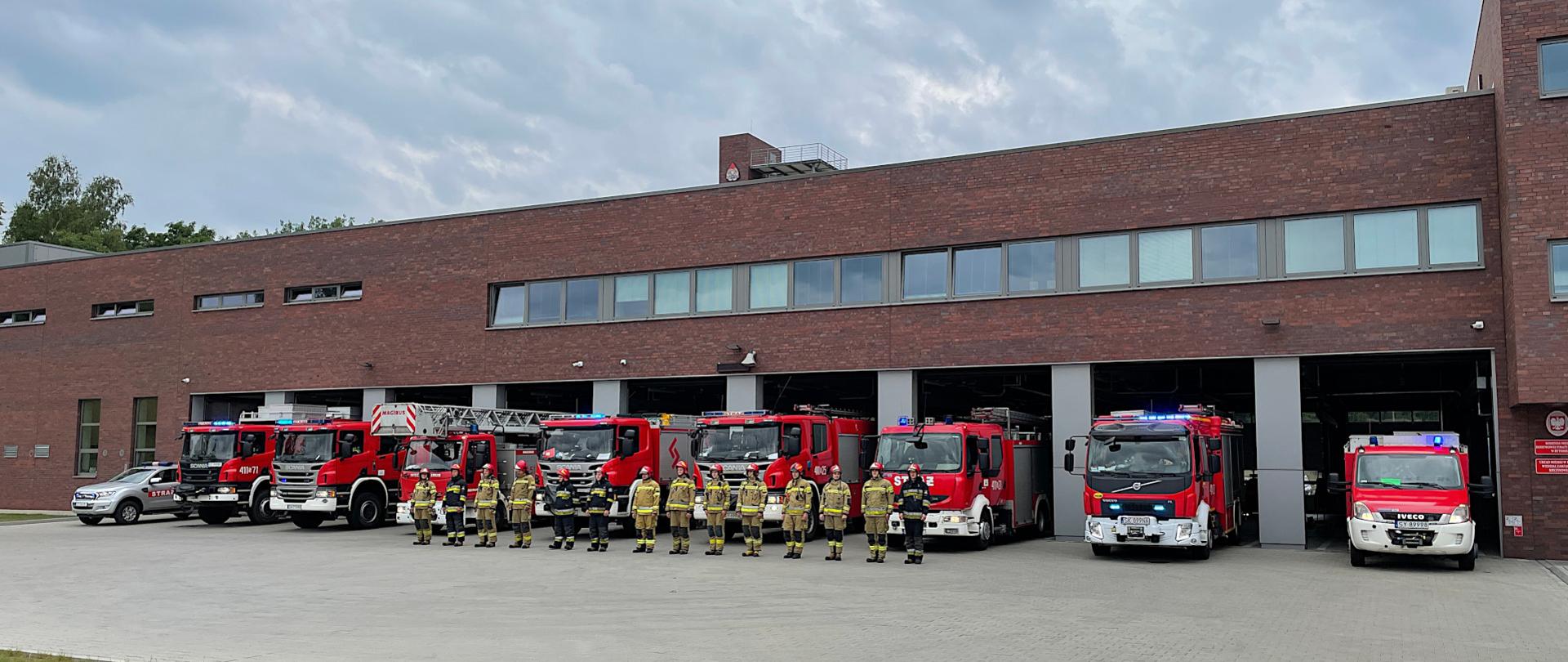 Na zdjęciu widoczny jest budynek komendy PSP. Przed otwartymi bramami garaży stoją samochody pożarnicze z włączonymi sygnałami dźwiękowymi i świetlnymi. Przed samochodami stoją, w szeregu strażacy w ubraniach specjalnych.