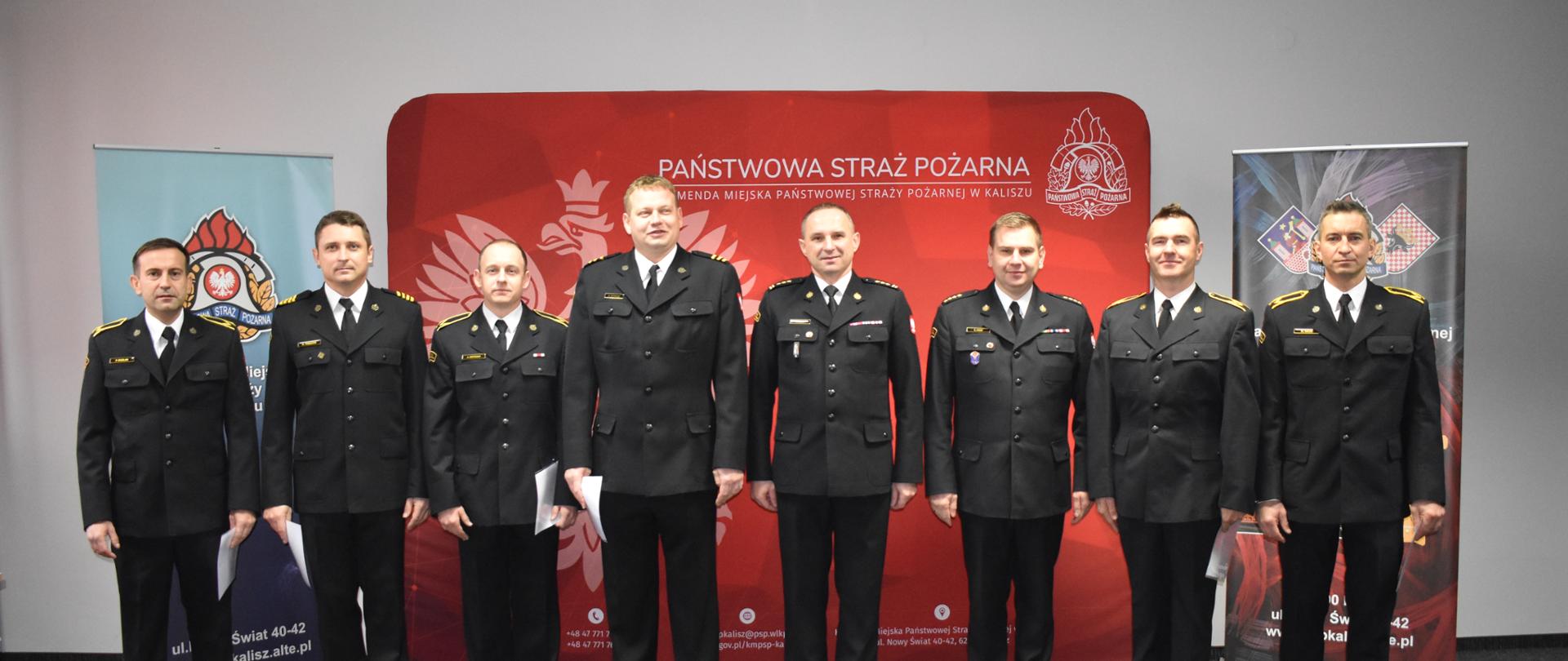 Na zdjęciu widać grupę wyróżnionych i awansowanych funkcjonariuszy\a KM PSP w Kaliszu. Za plecami ściany pomieszczenia. Na suficie lampy oraz kasetony w kolorze białym. Osoby stoją na szarym dywanie. Oświetlenie sztuczne, barwa zimna.