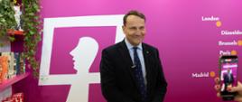 Wicepremier Sikorski w ciemnym garniturze i granatowym krawacie stoi przed różową ścianą z napisem „Polish Institutes – culture”. Obok znajdują się półki z książkami i zielone rośliny, a po prawej stronie ktoś fotografuje go smartfonem.