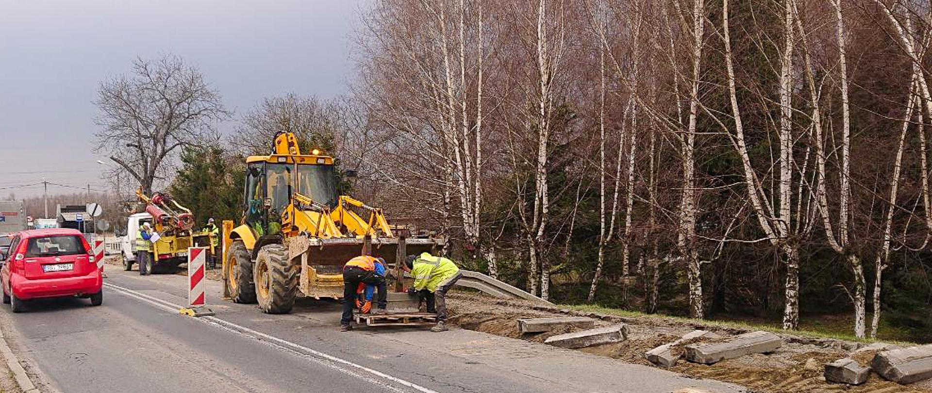 Fotografia z poziomu terenu prac budowlanych. Ruch drogowy na jedno jezdniowej drodze odbywa się lewym pasm. Na prawym pasie trwają prace rozbiórkowe. 