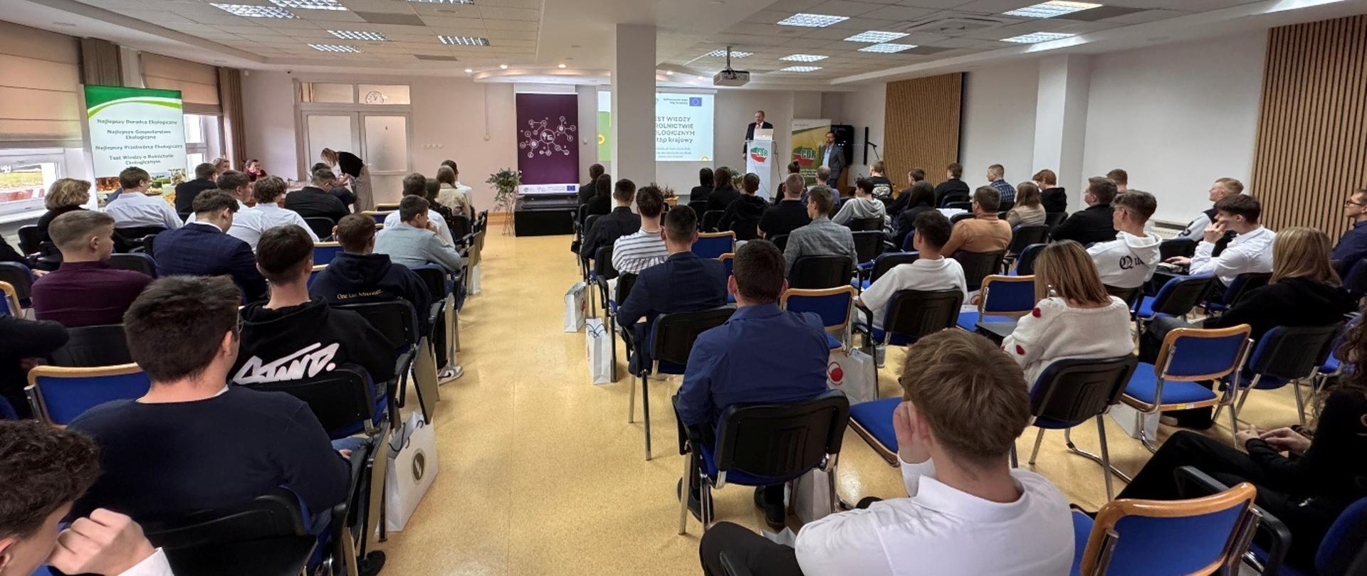 Szerokie ujęcie sali konferencyjnej, w której grupa kilkudziesięciu młodych osób (uczniów) siedzi na krzesłach, uczestnicząc w prelekcji. Prelegent stoi na niewielkiej scenie, a w tle widać banery promocyjne oraz prezentację wyświetlaną na ekranie.