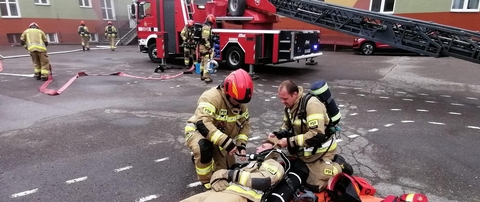 Strażacy ćwiczą na placu wewnętrznym szkoły. Na pomarańczowej macie leży strażak, udaje poszkodowanego, przy mim inni strażacy.W tle drabina mechaniczna strażacka i budynek szkoły.