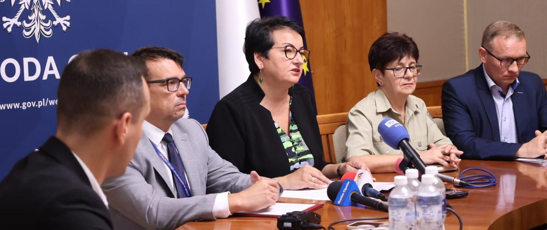 Wojewoda siedzi w Sali Złotej i mówi do mikrofonu. Wraz z nią siedzą dyrektor Wydziału Bezpieczeństwa i Zarządzania Kryzysowego, a także podległe służby. Wojewoda mówi do mikrofonów, które leżą na stole. Patrzy na wprost, w kierunku dziennikarzy.
