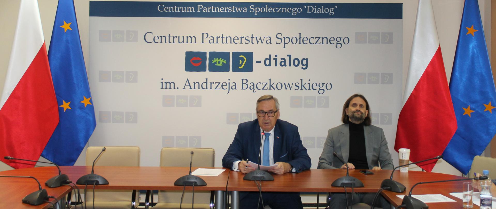 Zdjęcie przedstawia stół prezydialny za którym siedzą: Stanisław Szwed - Sekretarz Stanu w Ministerstwie Rodziny i Polityki Społecznej, Współprzewodniczący Zespołu ze strony rządowej, Piotr Bromber - Podsekretarz Stanu w Ministerstwie Zdrowia. W tle siedzących znajduje się baner z napisem Centrum Partnerstwa Społecznego Dialog i. Andrzeja Bączkowskiego. Po lewej i prawej stronie zdjęcia stoją po dwie flagi: Polska i UE. Nad banerem widnieje Godło Polski.