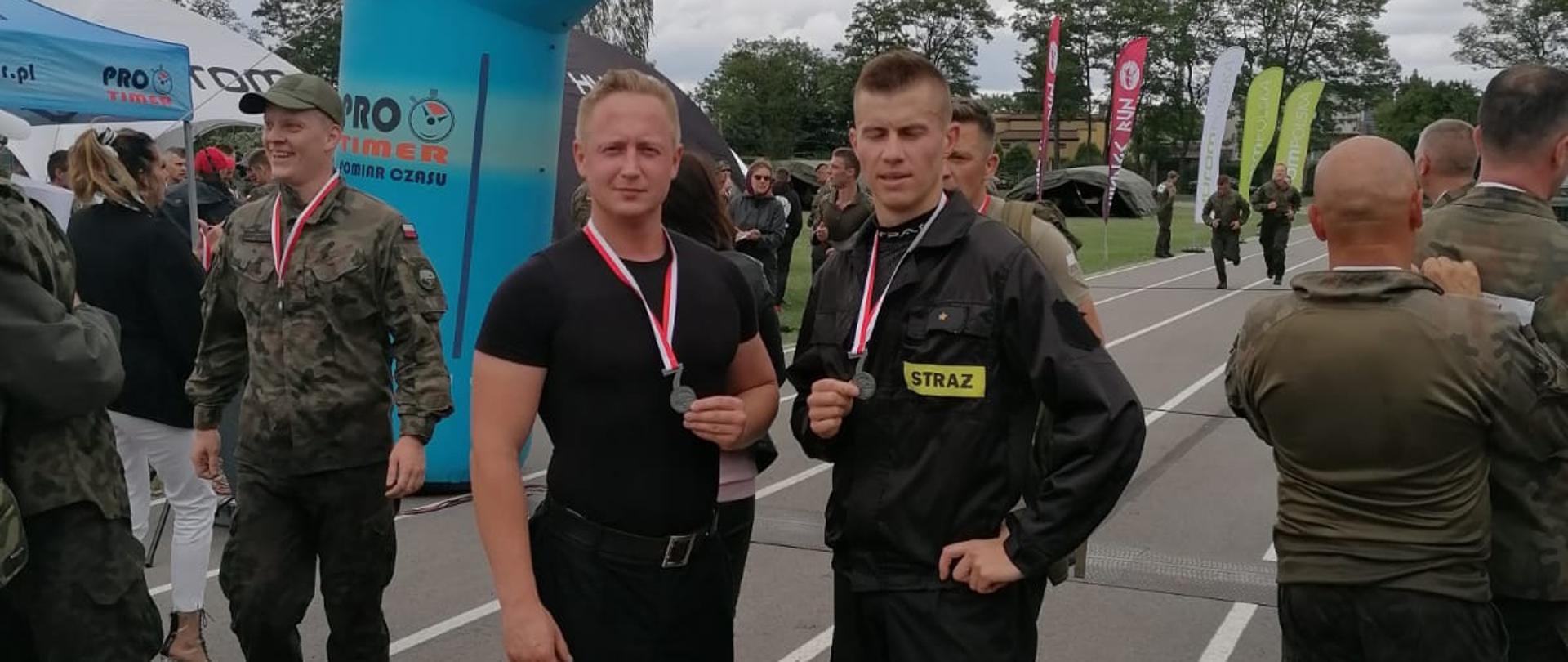 Na tle pochmurnego nieba duża dmuchana brama w kolorze niebieskim a na niej napis META. Przed bramą dwóch strażaków prezentuje zawieszone na szyj medale. Wokół nich znajdują się żołnierze. 