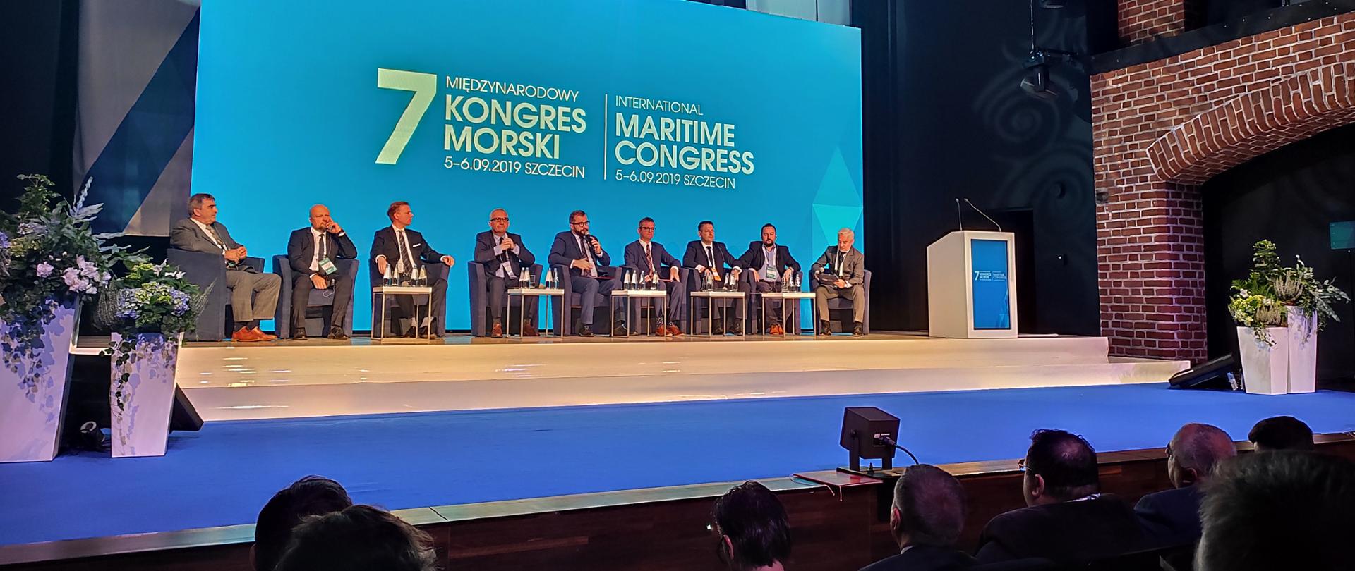 Zdjęcie z widokiem na scenę, na której siedzi dziewięciu mężczyzn, wśród nich minister Puda, który w ręku trzyma mikrofon. Za nimi wyświetlany jest napis 7 Międzynarodowy Kongres Morski w Szczecinie, 5-6.09.2019 Szczecin