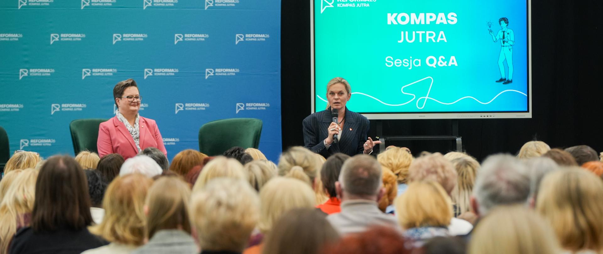 Minister Edukacji Barbara Nowacka przemawia do nauczycieli, uczestników spotkania "Kierunek: Kompas Jutra". Obok niej stoi wiceminister Katarzyna Lubnauer. Kobiety stoją na tle ścianki Kompasu Jutra, obok na telewizorze wyświetla się "Kompas Jutra - Sesja Q&A".