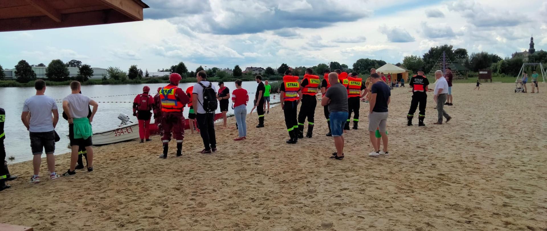  Na zdjęciu plaża i woda. Na plaży stoi wiele osób, wśród nich strażacy biorcy udział w ćwiczeniach z ratownictwa wodnego.