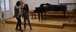 Na zdjęciu widnieje nauczyciel fortepianu mgr Dorota Skibicka, która w auli szkoły tańczy wraz z uczniem. Tło zdjęcia jest białe i jasno brązowe, w tle czarny fortepian.