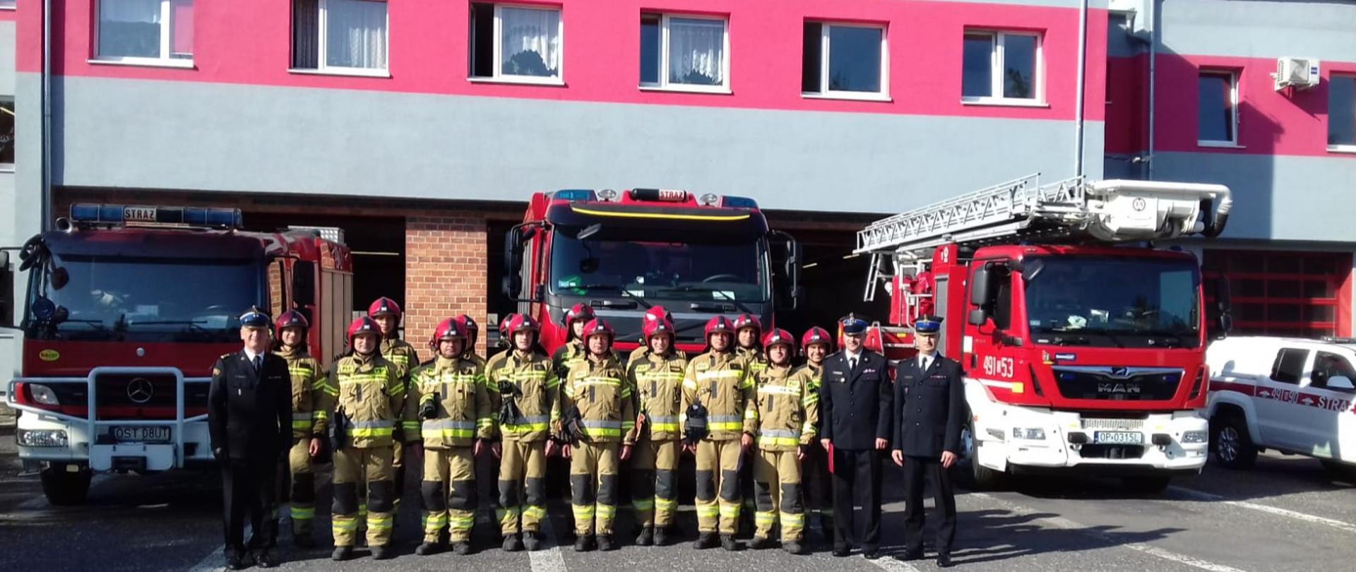 Na zdjęciu znajdują się strażacy w mundurach na tle samochodów pożarniczych i budynku komendy