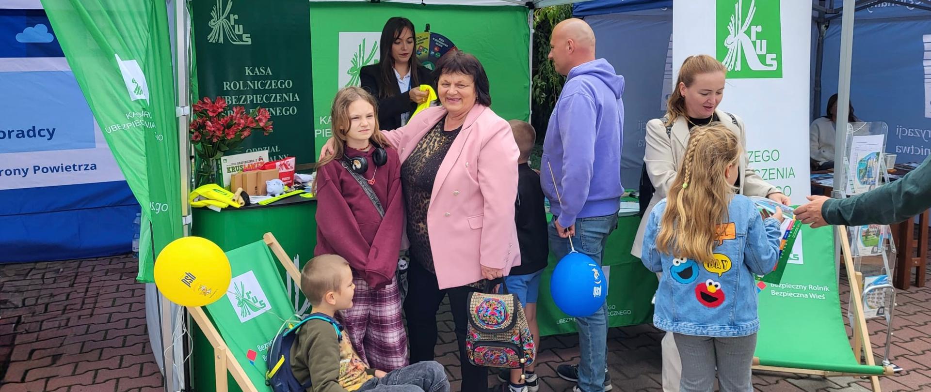 Biało-zielony namiot z logotypami KRUS jest ustawiony na zewnątrz. Przed stoiskiem stoi kilka osób – dzieci i dorośli – rozmawiają, oglądają materiały promocyjne oraz biorą udział w wydarzeniu.