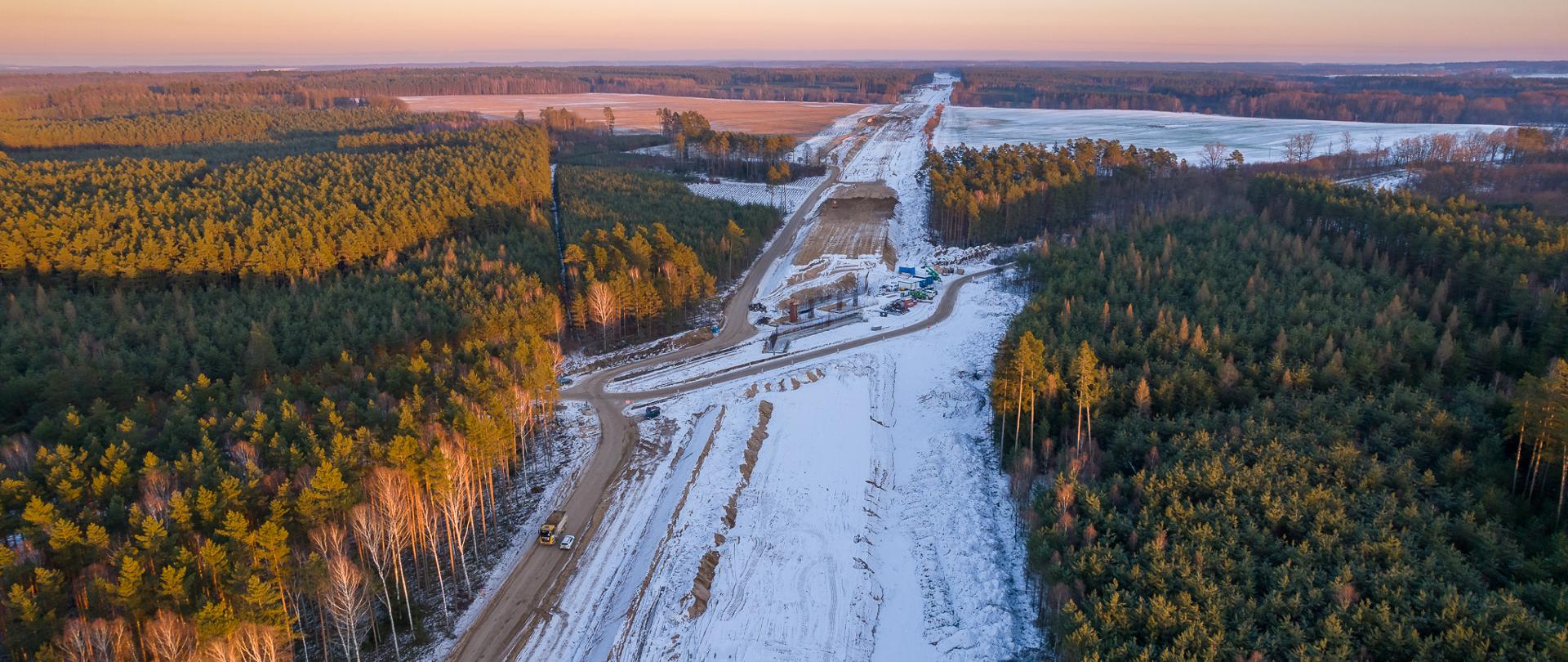 Na zdjęciu widoczny jest plac budowy drogi ekspresowej, otoczony lasem. Droga jest w trakcie budowy, z wyraźnie widocznymi pasami ziemi i śniegu. W tle można dostrzec maszyny budowlane oraz materiały potrzebne do kontynuacji prac. Cała scena rozciąga się na dużą odległość, ukazując postęp prac na różnych etapach budowy.