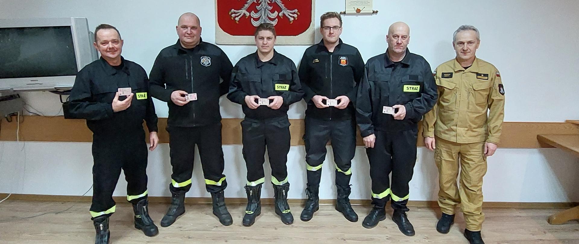 Kolorowa fotografia wykonana w pomieszczeniu, przedstawia pięciu strażaków ochotniczych straży pożarnych stojących strażaków w ubraniach koszarowych i jednego strażaka Państwowej Straży Pożarnej w jasnym ubraniu służbowym. Z tyłu na ścianie wiszące Godło Polski i obrazy.