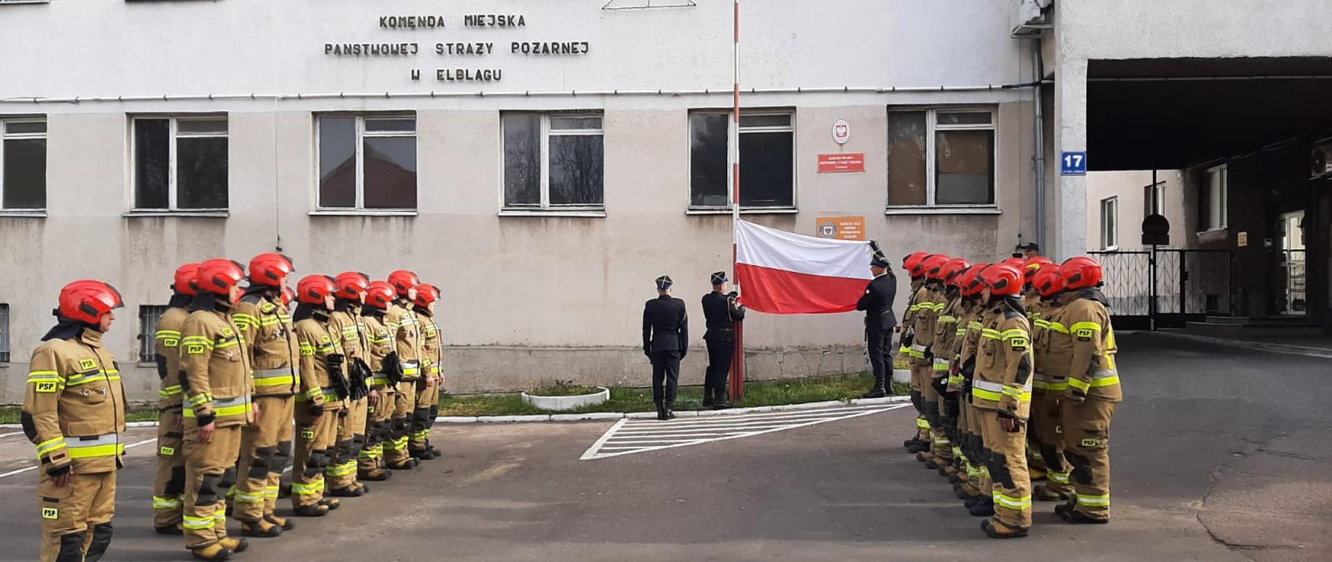 Poczet flagowy prezentuje flagę państwową. Po obu stronach stoją w szeregu strażacy.
