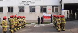Poczet flagowy prezentuje flagę państwową. Po obu stronach stoją w szeregu strażacy.