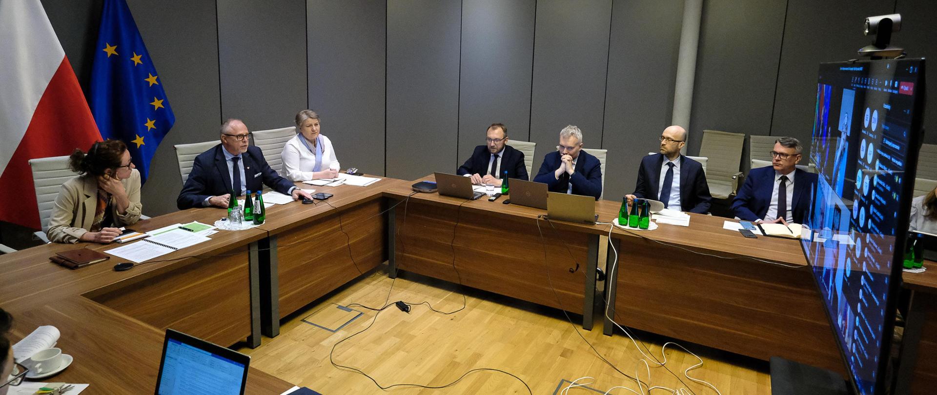 W sali konferencyjnej grupa osób siedzi przy stołach. Wśród nich wiceminister Jacek Protas. Za nim flagi PL i UE. Na środku stoi monitor.
