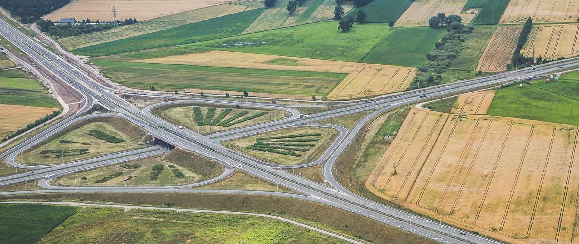 Widok z lotu ptaka na węzeł drogowy w Ostrowie Wielkopolskim. Wielopoziomowe skrzyżowanie dróg ekspresowych z łącznicami i estakadami otoczone jest polami uprawnymi w różnych odcieniach zieleni i złota. Na jezdniach widoczny jest niewielki ruch samochodowy.