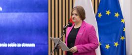 Zdjęcie z boku, na tle ściany z pionowych drewnianych listewek z wbudowanym wielkim ekranem stoi wiceminister Zioło-Pużuk w różowej marynarce i mówi do mikrofonu na stojaku, za nią flagi Polski i UE.