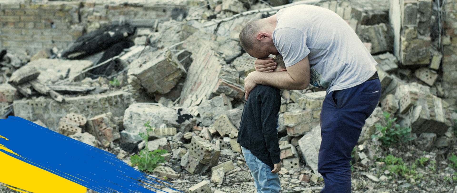 W lewym dolnym rogu flaga Ukrainy, w środku kawałki zburzonego budynku i mężczyzna, który przytula małego chłopca.