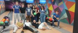 To zdjęcie przedstawia grupę osób spędzających wspólnie czas na kręgielni.
Wielu członków grupy ma na sobie odzież sportową, w tym koszulki z logo ""Kujavia"", co sugeruje przynależność do klubu sportowego z Włocławka.
Na zdjęciu widoczne jest również logo marki Adidas na jednej z koszulek.
Uczestnicy znajdują się na profesjonalnym torze bowlingowym, co jest popularną formą integracji sportowej wśród grup szkolnych i sportowych.
W tle widoczne są ekrany wyświetlające wyniki gry, co potwierdza, że jest to aktywne spotkanie rekreacyjne.