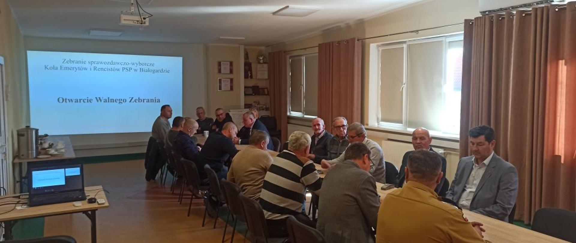 Zdjęcie wykonane z głębi sali w stronę ekranu projekcyjnego, na którym wyświetlany jest slajd z napisem: „Zebranie sprawozdawczo-wyborcze Koła Emerytów i Rencistów PSP w Białogardzie" oraz nagłówek „Otwarcie Walnego Zebrania". Uczestnicy siedzą przy stołach, zwróceni w stronę ekranu. Na pierwszym planie po lewej stronie widać laptopa sterującego prezentacją.