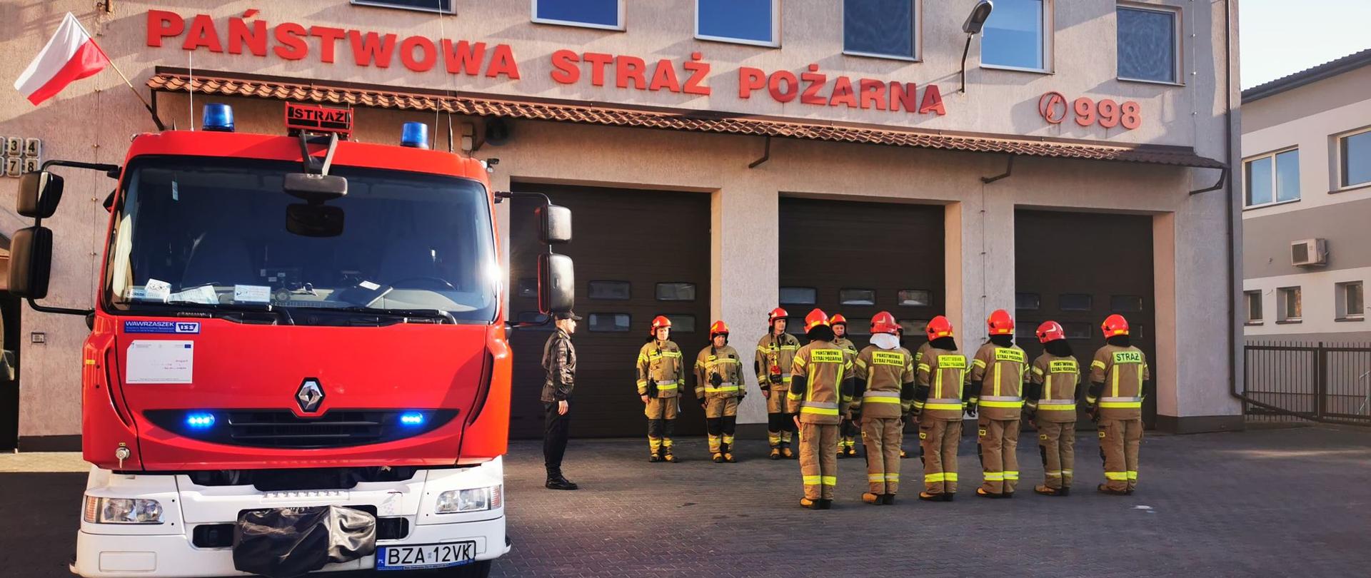 Zdjęcie przedstawia strażaków w ubraniu bojowym, stojących w szeregu na przeciw siebie. Z lewej stronie dowódca Jednostki Ratowniczo-Gaśniczej stoi ubrany w ubranie dowódczo-sztabowe. Z lewej strony zdjęcia znajduje się samochód ratowniczo-gaśniczy. Zdjęcie wykonano w słoneczny dzień na placu Komendy.