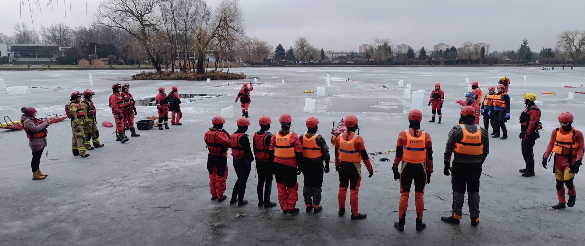 Zdjęcie przedstawia grupę ratowników biorących udział w ćwiczeniach na zamarzniętym zbiorniku wodnym. Na pierwszym planie widoczny jest rząd osób tyłem do obiektywu, ubranych w profesjonalne, jaskrawe kombinezony ratunkowe oraz kamizelki z napisem WOPR (Wodne Ochotnicze Pogotowie Ratunkowe). Wszyscy uczestnicy mają na głowach kaski ochronne, co podkreśla szkoleniowy i bezpieczny charakter operacji.
W tle widać rozległą, pokrytą lodem przestrzeń z wyciętymi przeręblami, przy których pracują pozostali ratownicy oraz strażacy w piaskowych mundurach. Na lodzie znajduje się specjalistyczny sprzęt, taki jak sanie lodowe i liny. Otoczenie jeziora stanowią nagie drzewa, niska zabudowa oraz pochmurne, zimowe niebo, co tworzy surową, lecz profesjonalną atmosferę przygotowań do ratownictwa lodowego.
