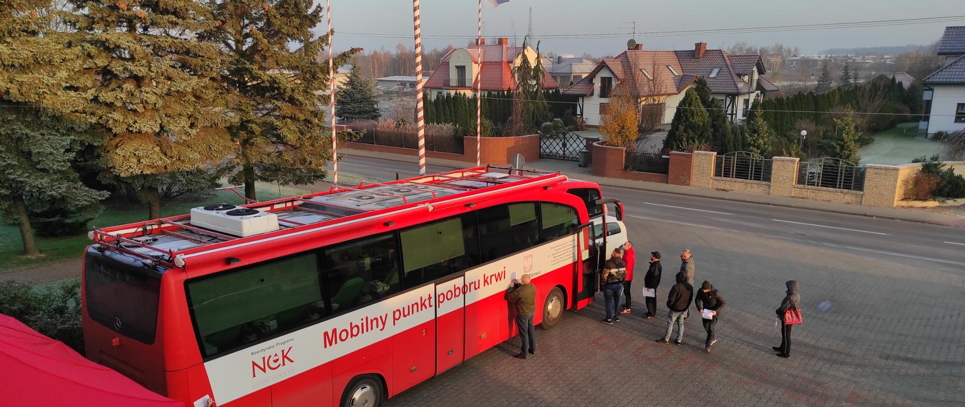 Zdjęcie przedstawia akcję krwiodawstwa organizowaną prze KP PSP w Koluszkach w dniu 26.11.2020 r. Na zdjęciu widoczny jest autobus krwiodawstwa z napisem "Mobilny punkt poboru krwi" oraz kolejka chętnych do oddania krwi. Zdjęcie przed budynkiem KP PSP w Koluszkach. 