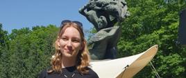 Na tle pomnika Fryderyka Chopina w warszawskich Łazienkach stoi młoda laureatka konkursu z dyplomem w dłoniach.