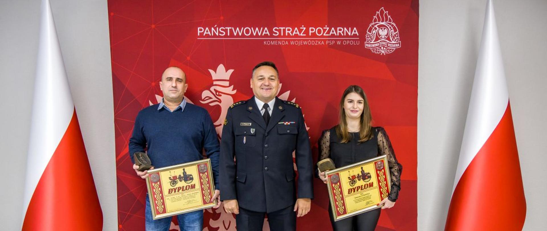 Wspólne zdjęcie Opolskiego Komendanta Wojewódzkiego PSP w Opolu st. bryg. Krzysztofa Kędryka, Pani Katarzyny Szymańskiej oraz asp. sztab. w st. spoczynku Rafała Sajkowskiego na tle banneru PSP i flag RP.