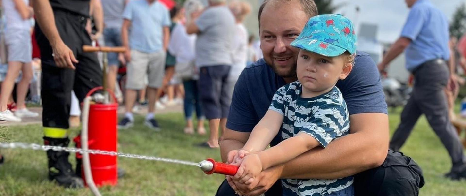 Na pierwszym planie dziecko z tatą leje wodę z trzymanej prądownicy. W tle strażak z czerwoną hydronetką.