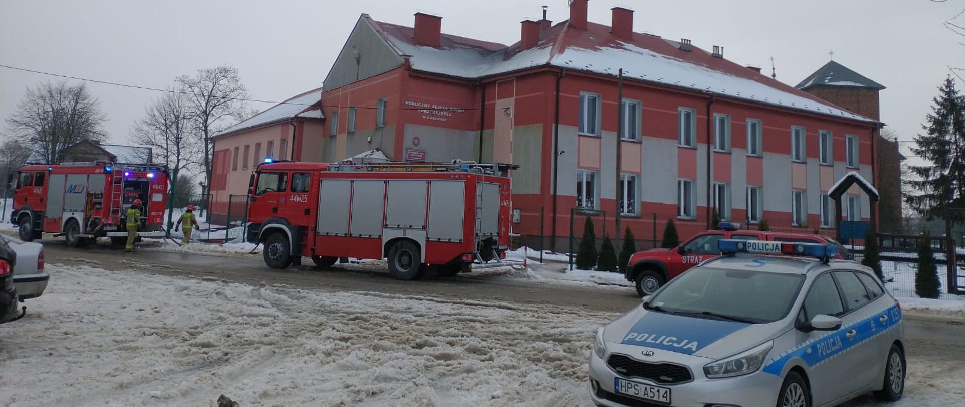 Zdjęcie przedstawia trzy czerwone samochody Jednostek Ochrony Przeciwpożarowej oraz jeden policyjny radiowóz. Samochody stoją na drodze naprzeciwko Publicznej Szkoły Podstawowej w Lasocinie. W tle widać budynek Publicznej Szkoły Podstawowej w Lasocinie.