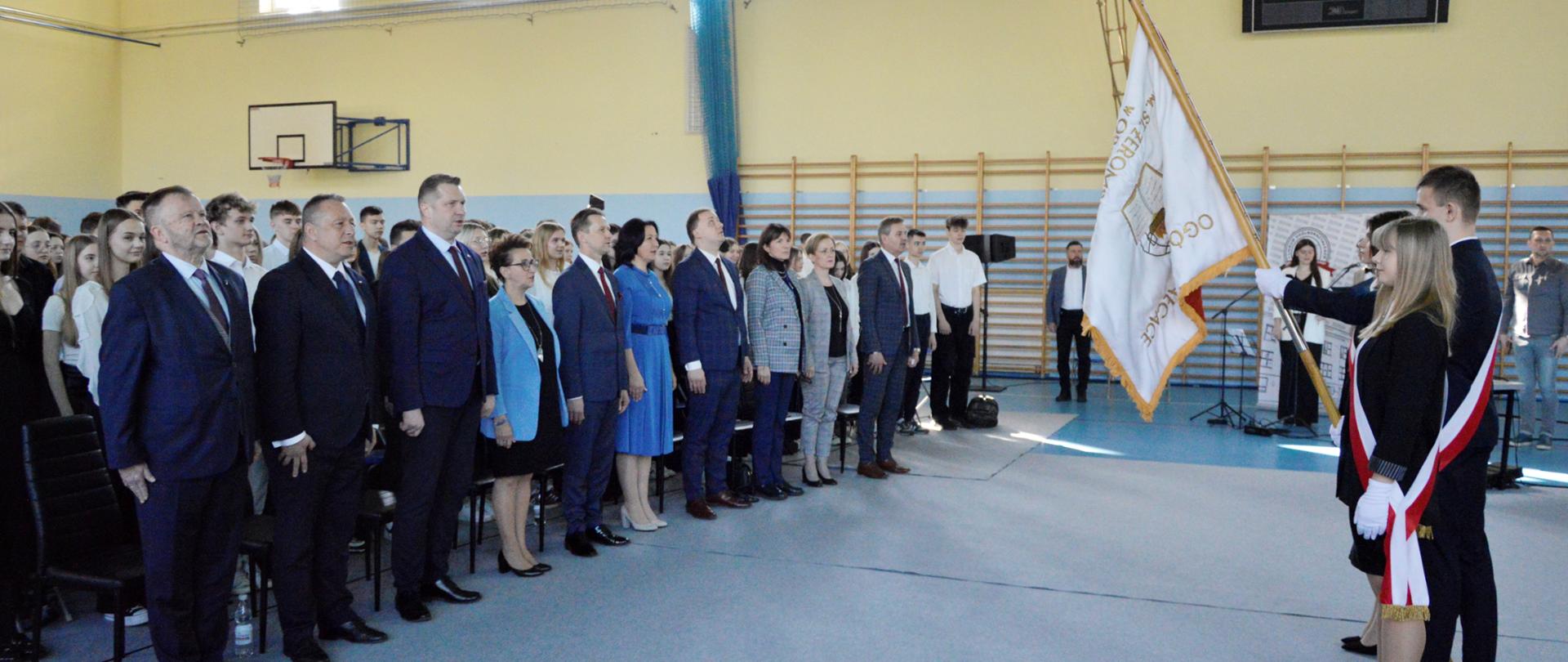 Widok z boku na salę gimnastyczną, po lewej duża grupa ludzi stoi w równych rzędach, po prawej troje młodych elegancko ubranych ludzi przepasanych biało-czerwonymi wstążkami, chłopak pośrodku trzyma biało-czerwony sztandar ze złotymi frędzlami.