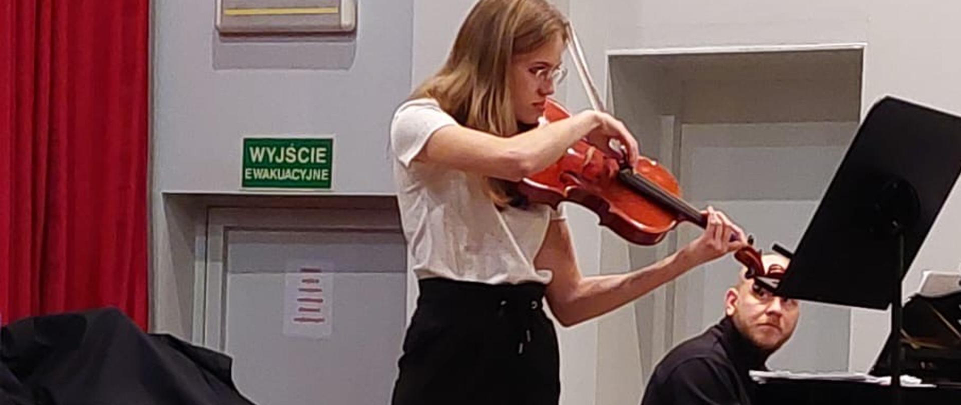 Nastolatka stojąc na estradzie auli gra na altówce, za nią na fortepianie gra mężczyzna.