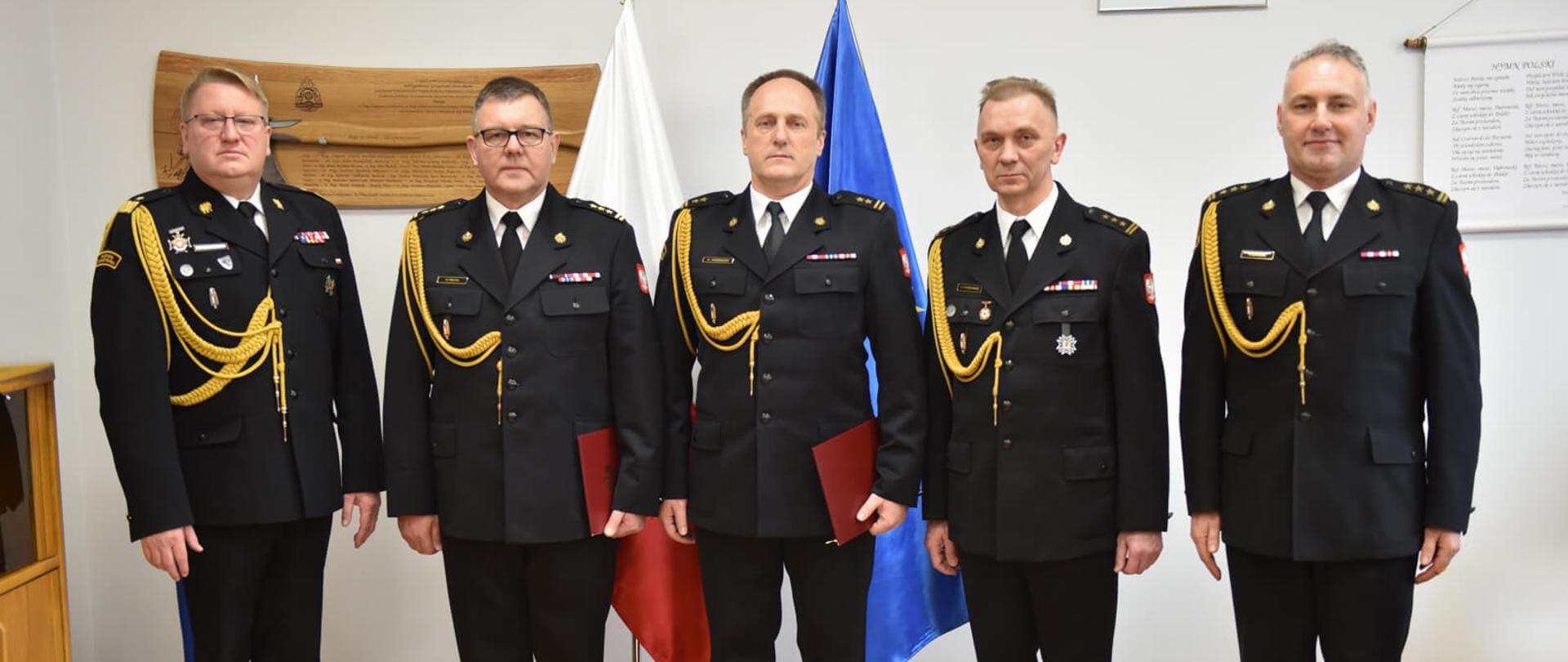 pięciu strażaków w mundurach galowych pozuje do zdjęcia na tle flagi polski i unii europejskiej
