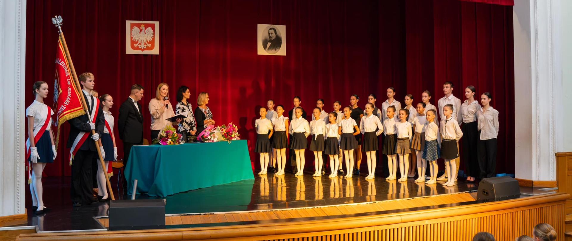 Zdjęcia z Dnia Edukacji Narodowej, aula szkoły. Po lewej stronie poczet, na środku dyrekcja po lewej nowi uczniowie ślubujący. W tle zdjęcie patrona oraz godło szkoły na czerwonym horyzoncie za sceną.