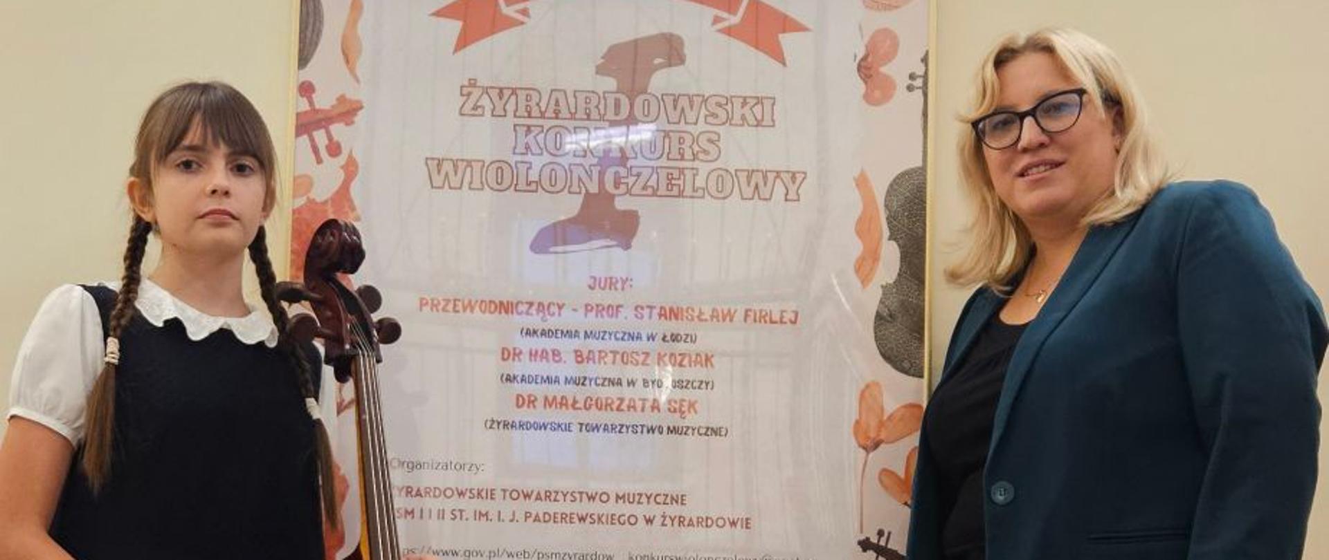 Zdjęcie przestawia Weronikę Krzyżewską i panią Urszulę Właźniak stojące po bokach plakatu reklamującego I Żyrardowski Konkurs Wiolonczelowy. Plakat w kolorach: brązowo, pomarańczowo, czarno, szarym. Na górze plakatu wiolonczela, poniżej informacje dotyczące konkursu. Nad plakatem okno. 