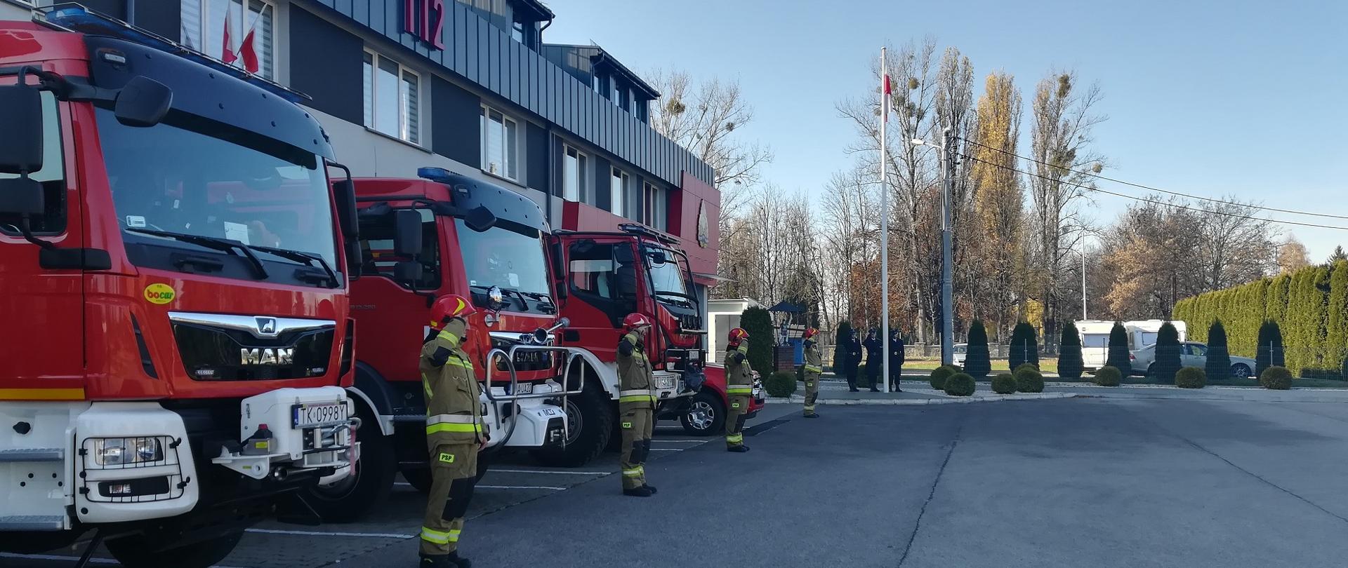 Zdjęcie przedstawia strażaków stojących w szeregu przed samochodami pożarniczymi oddających honor z okazji podniesienia flagi państwowej na maszt.