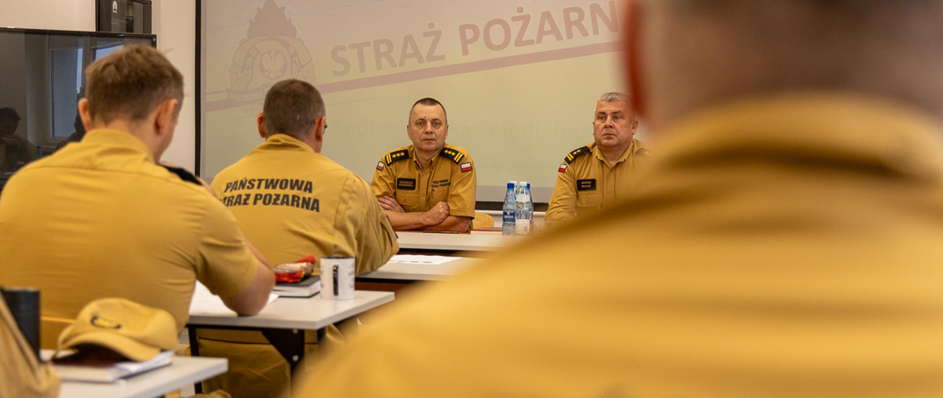 Uczestnicy warsztatów siedzą w sali konferencyjnej, zwróceni w stronę dwóch prowadzących siedzących przy stole prezydialnym. Za nimi widoczny jest ekran z logotypem PSP i napisem „Państwowa Straż Pożarna”. Zdjęcie przedstawia część teoretyczną warsztatów, dotyczącą m.in. dokumentacji, uprawnień KDR oraz współpracy między jednostkami.