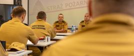 Uczestnicy warsztatów siedzą w sali konferencyjnej, zwróceni w stronę dwóch prowadzących siedzących przy stole prezydialnym. Za nimi widoczny jest ekran z logotypem PSP i napisem „Państwowa Straż Pożarna”. Zdjęcie przedstawia część teoretyczną warsztatów, dotyczącą m.in. dokumentacji, uprawnień KDR oraz współpracy między jednostkami.
