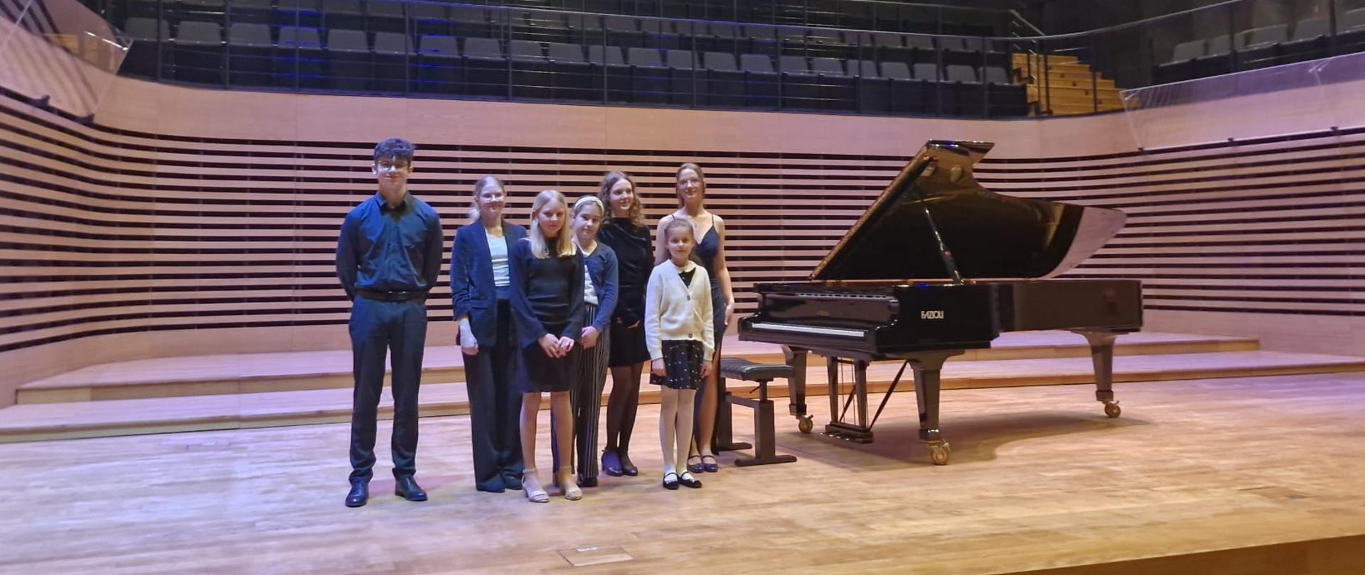 Sześć dziewczyn i jeden chłopka stoją na estradzie przy fortepianie.