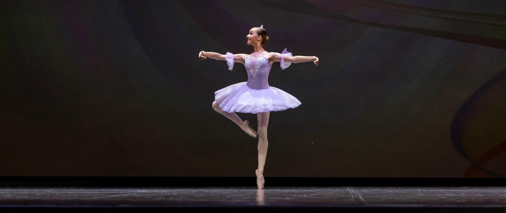 Zdjęcie Zuzanny Boryczki na scenie, uczennica w pozie baletowej ubrana w liliową paczkę klasyczną. Czarne tło. 