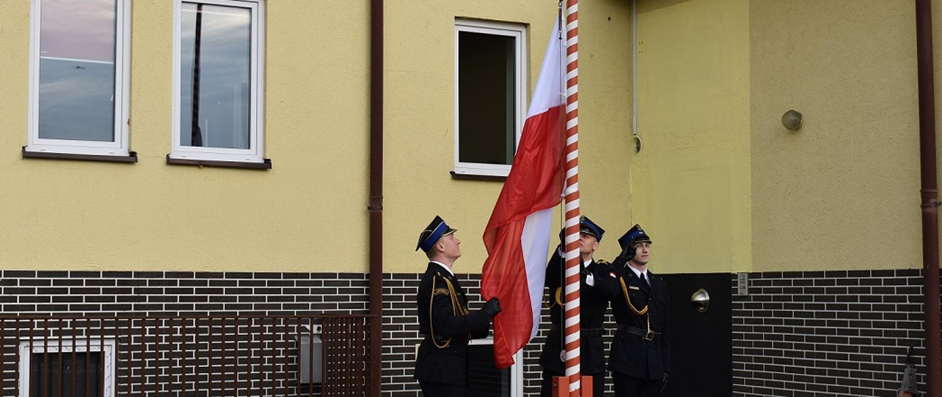 Dzień. Widok na ścianę budynku w kolorze żółtym. Przed budynkiem stoi maszt flagowy. Trzech funkcjonariuszy ubranych w umundurowanie wyjściowe, złote sznury galowe oraz rogatywki stoi przy maszcie. Jednej z nich trzyma flagę Polski. Drugi wciąga flagę Polski na maszt. Trzeci stoi obok oddając honory - salutuje.
